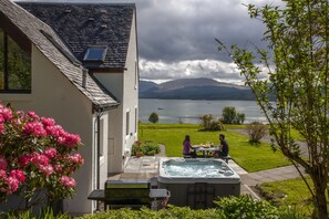 A house with a hot tub, a dining table, and a view of a lake and mountains. - Large cottage on shore of Scottish Loch (Benderloch)