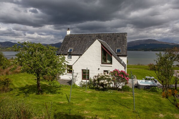 A white house with a sloped roof, a hot tub, and a view of a lake and mountains. - Large cottage on shore of Scottish Loch (Benderloch)