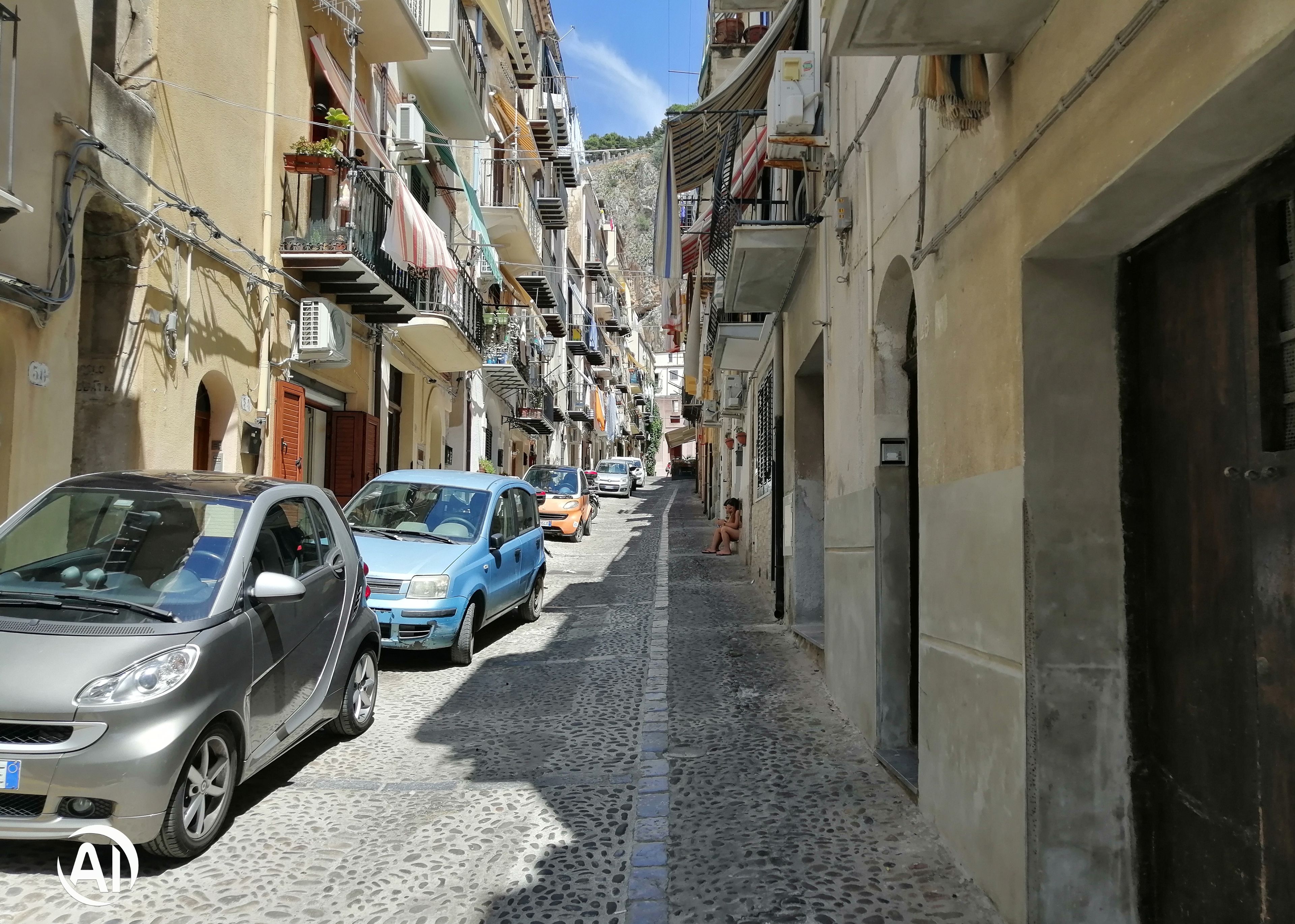 A narrow cobblestone street lined with multi-story buildings, parked cars, and balconies with plants and laundry.