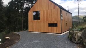 A wooden cabin with a sloped roof, a gravel driveway, and a forested backdrop. - Red Stag With Stunning Loch and Waterfall View (Crianlarich)