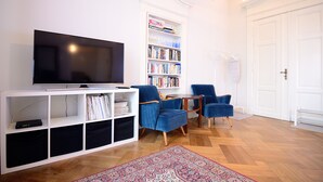 A living room with a flat-screen TV, a bookshelf, two blue armchairs, a coffee table, and a patterned rug. - Victus Apartamenty - Bolek (Sopot)