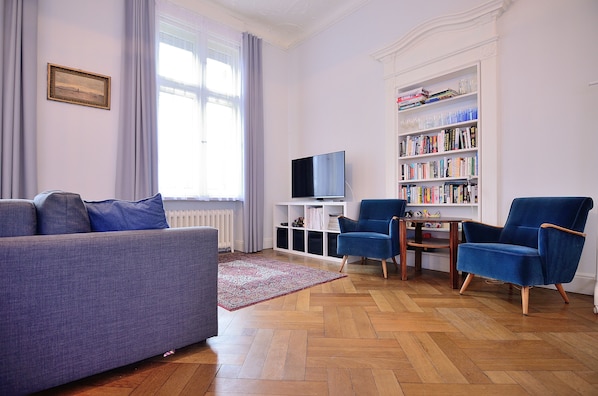 A living room with a sofa, two armchairs, a TV, a bookshelf, and a window with curtains.