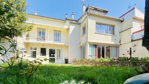 Une maison à deux étages avec un balcon, un jardin fleuri et un ciel d’un bleu éclatant.