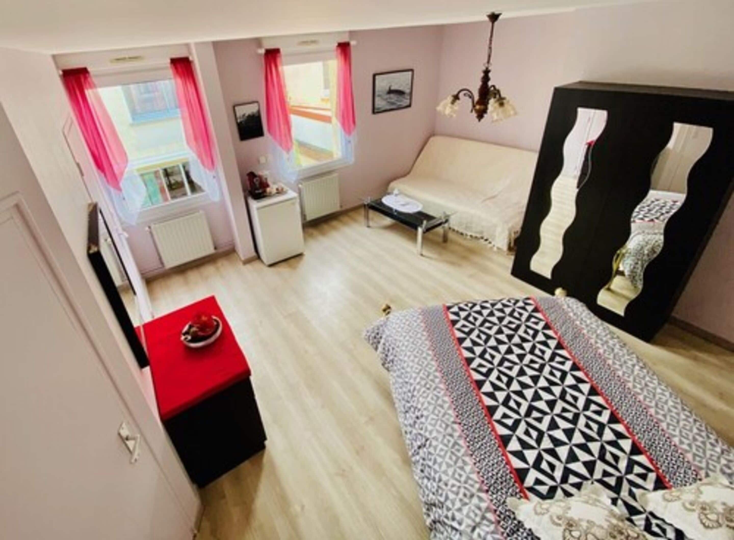 A bedroom with a bed, a red table, a white sofa, a black wardrobe, and a window with pink curtains.
