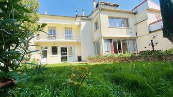 A two-story house with a balcony, a garden with flowers, and a clear blue sky.