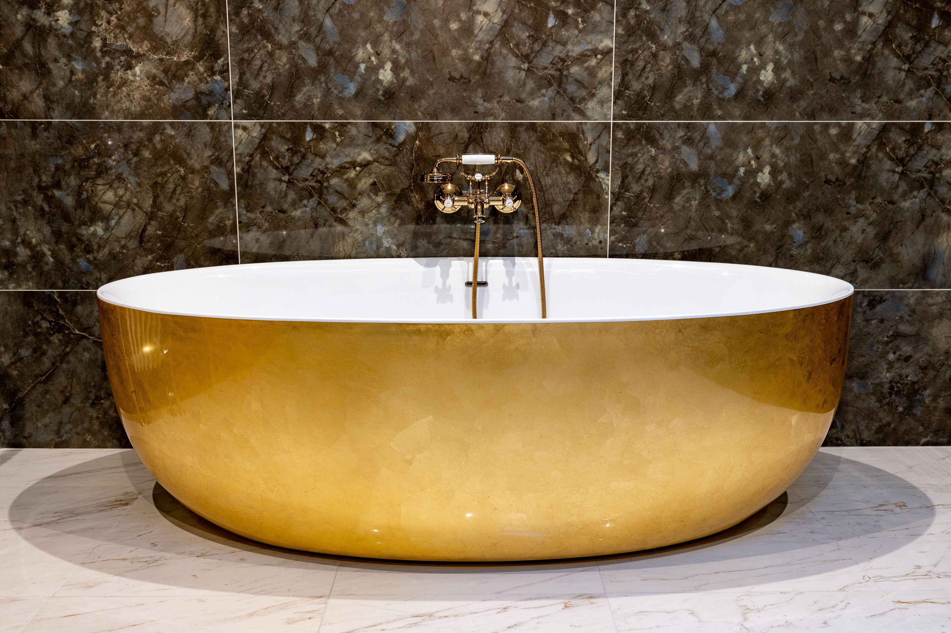 A large, oval-shaped bathtub with a gold finish and a white interior, set against a dark marble wall.
