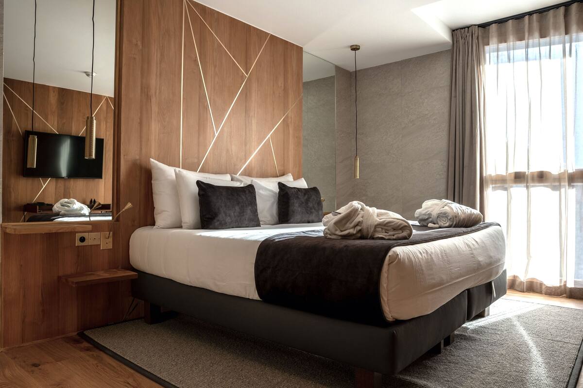 A modern hotel room with a large bed, a flat-screen TV, wooden paneling, and a view of the outdoors.