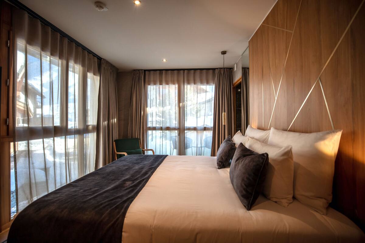 A modern hotel room with a large bed, a view of the outdoors, and a seating area with a chair.