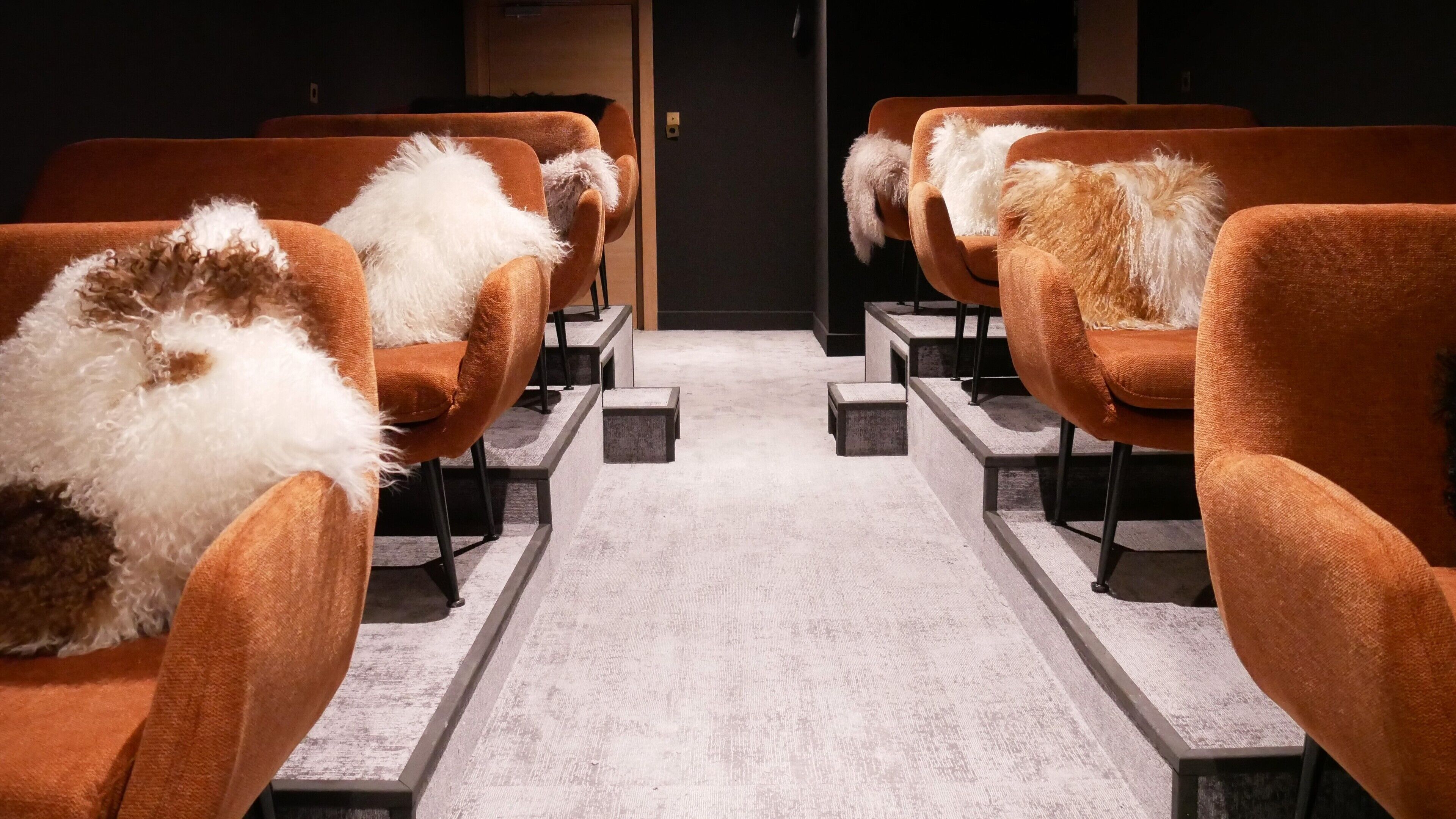Une rangée de chaises orange, agrémentées de coussins en fausse fourrure blanche, est disposée de manière linéaire. Le sol est recouvert d’une moquette grise et une porte en bois se trouve au fond.