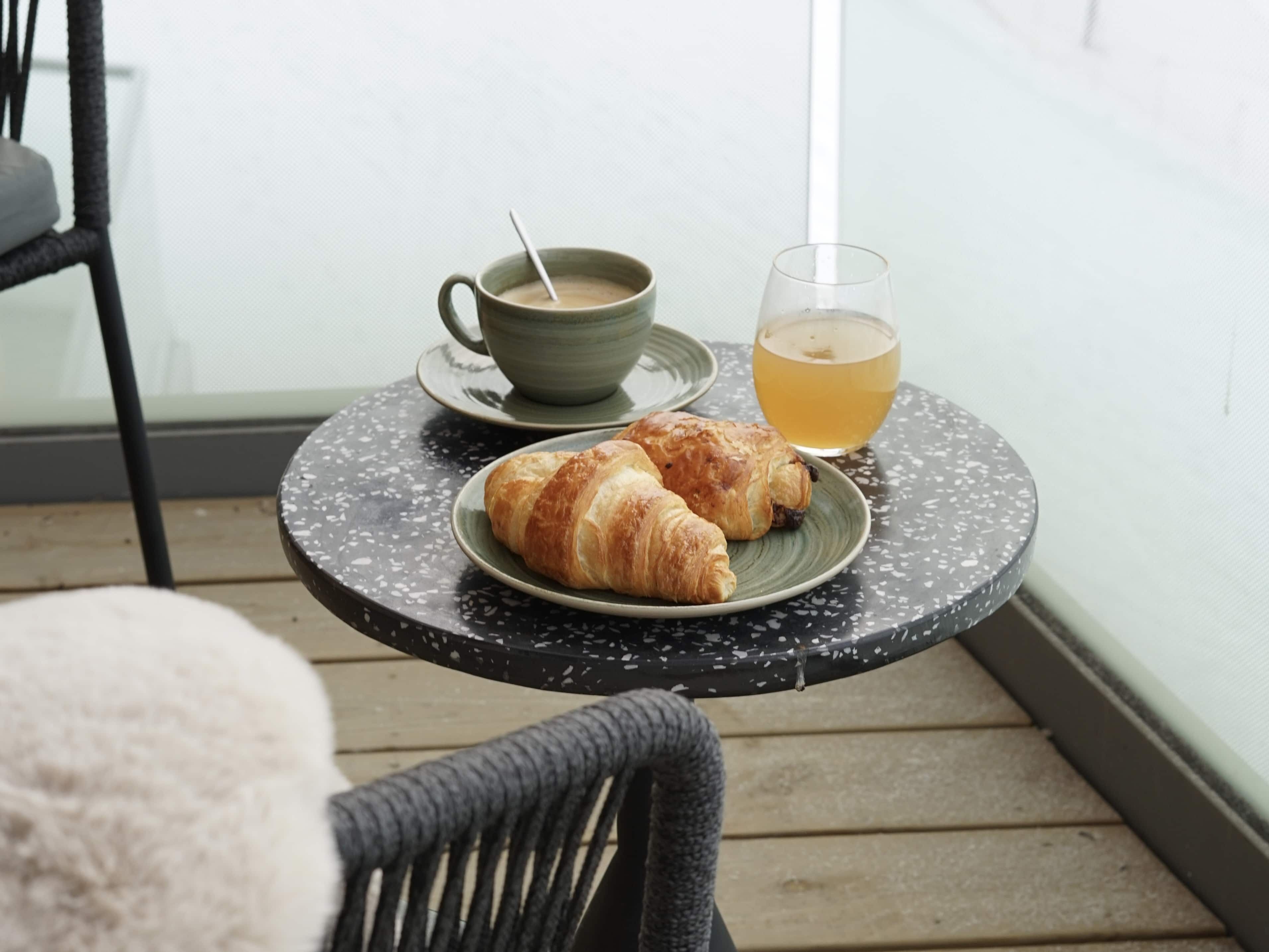 Et lille rundt bord med et fad croissanter, en kop kaffe og et glas te på en balkon.