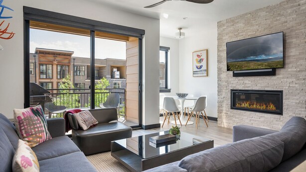 A modern living room with a fireplace, a large TV mounted on a brick wall, and a balcony with a view of the city.