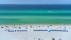 An aerial view of a beach with rows of blue and red umbrellas, a clear blue sky, and the ocean in the background.