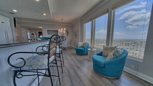 A spacious balcony with a view of the city and ocean, a dining area with a bar, and a living area with two blue armchairs and a coffee table.