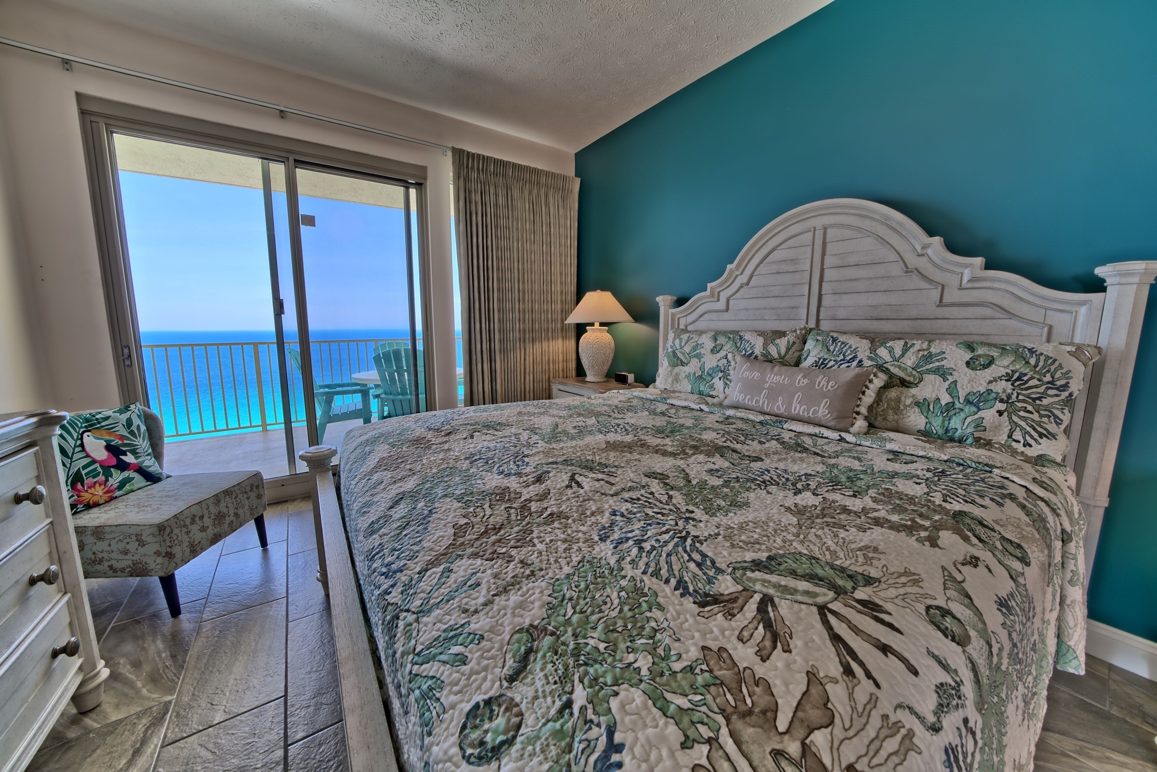 A bedroom with a large bed, a nightstand, a chair, and a view of the ocean through a sliding door.
