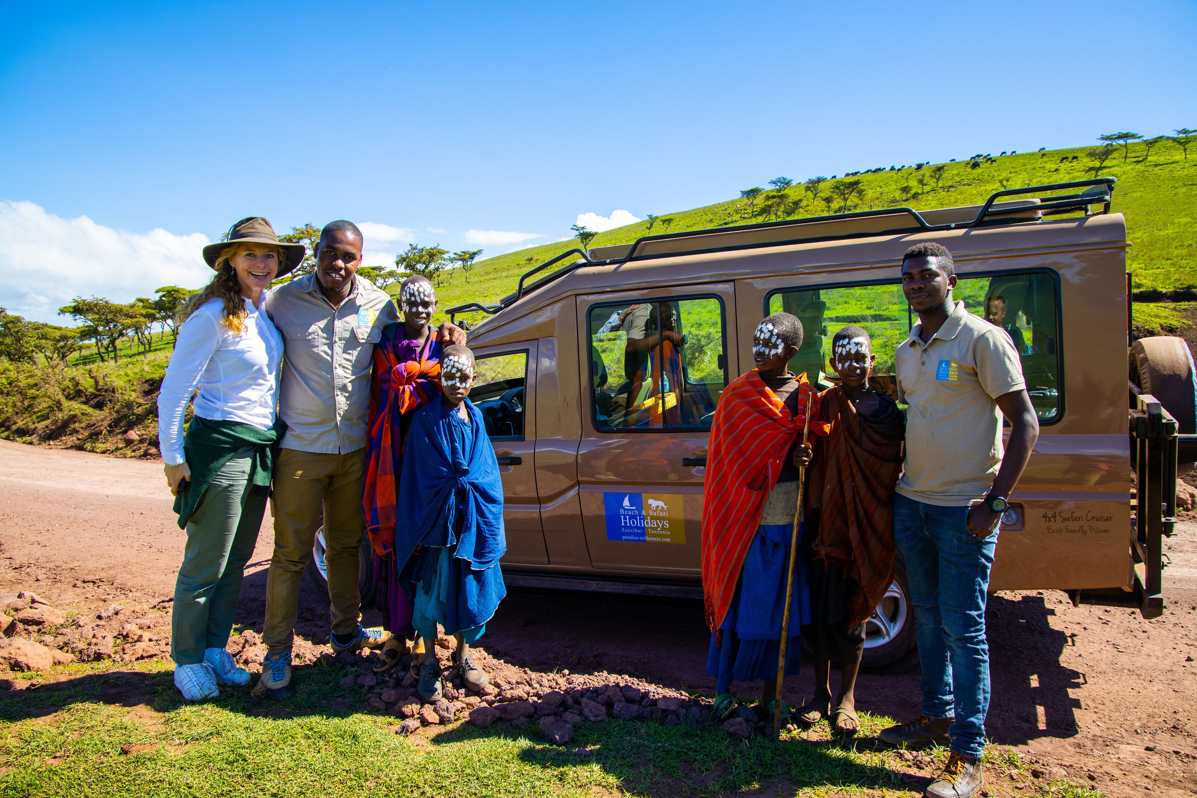 A group of people standing in front of a safari vehicle with a grassy landscape in the background.