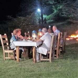 A group of people dining outdoors at night with a table set for a meal, surrounded by chairs, and a fire in the background. - Africa Safari Serengeti Ikoma (Serengeti)