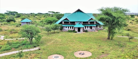 A large lodge with a green roof and multiple rooms, surrounded by a grassy landscape with trees and a dirt path.