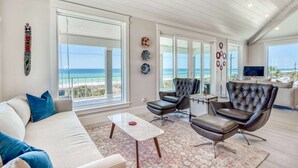 Een ruime woonkamer met een groot raam met uitzicht op het strand, leren fauteuils, een witte bank en een salontafel.