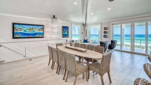 Een ruime eethoek met een houten tafel en geweven stoelen, grote ramen met uitzicht op de oceaan en een plafondventilator.