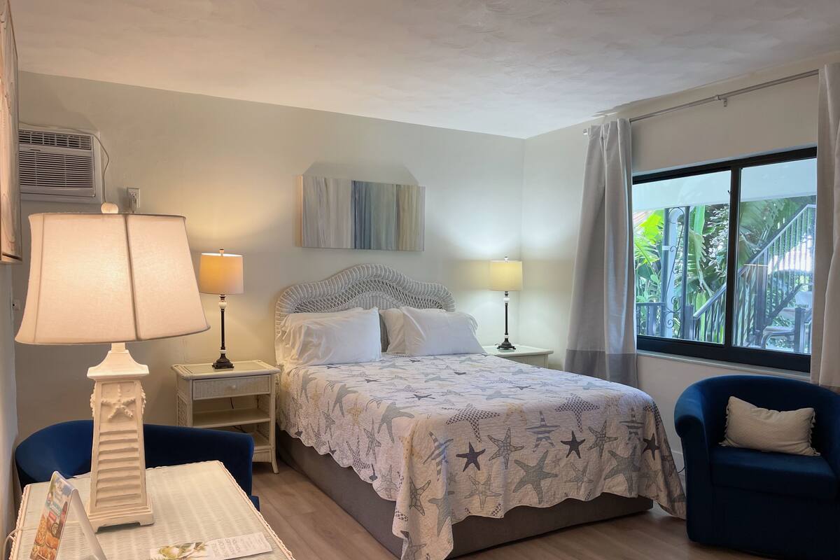 A hotel room with a bed, two bedside tables with lamps, a chair, a small table with a menu, and a window with a view of greenery.