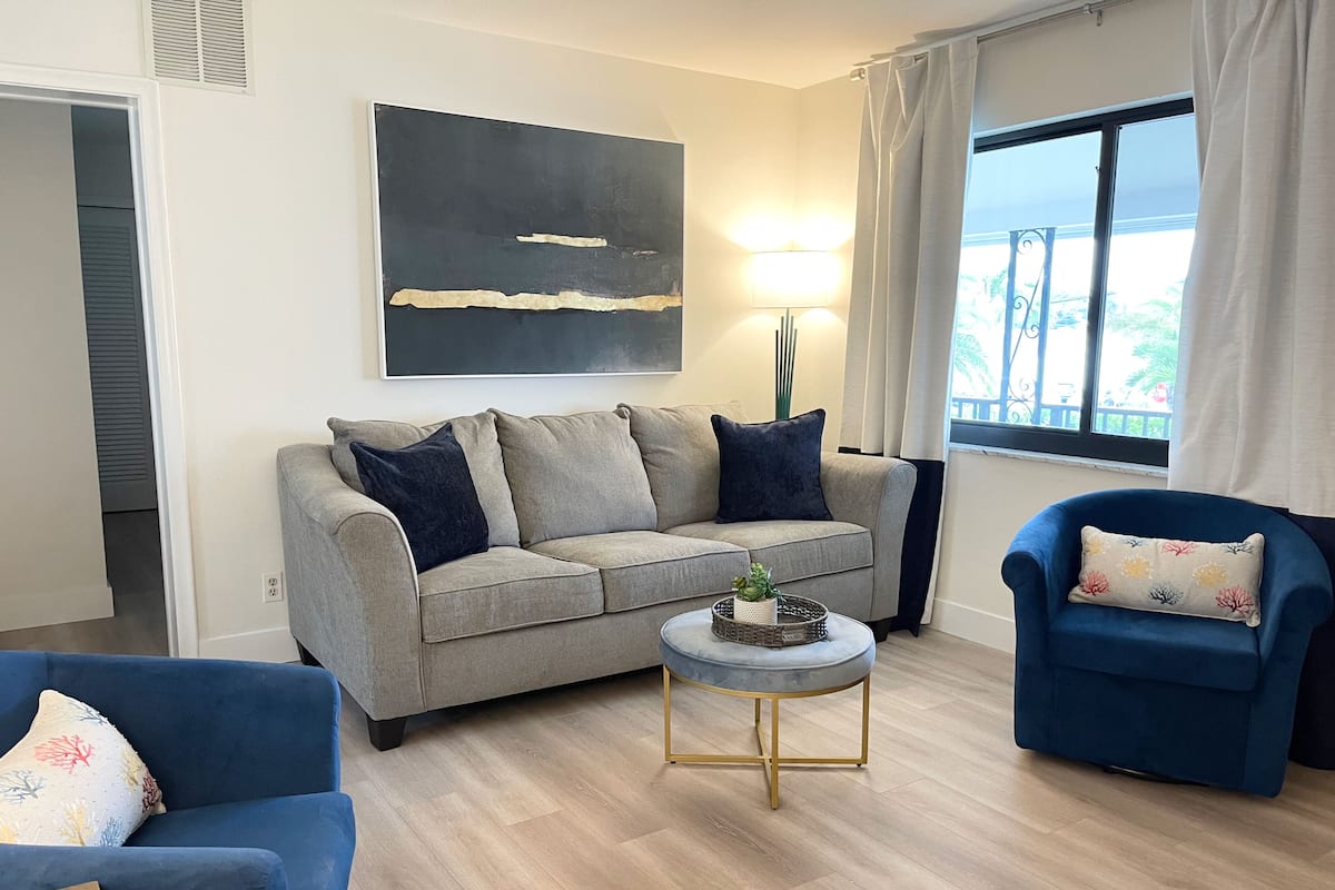 A living room with a grey sofa, a blue armchair, a small round table, and a large window with curtains.