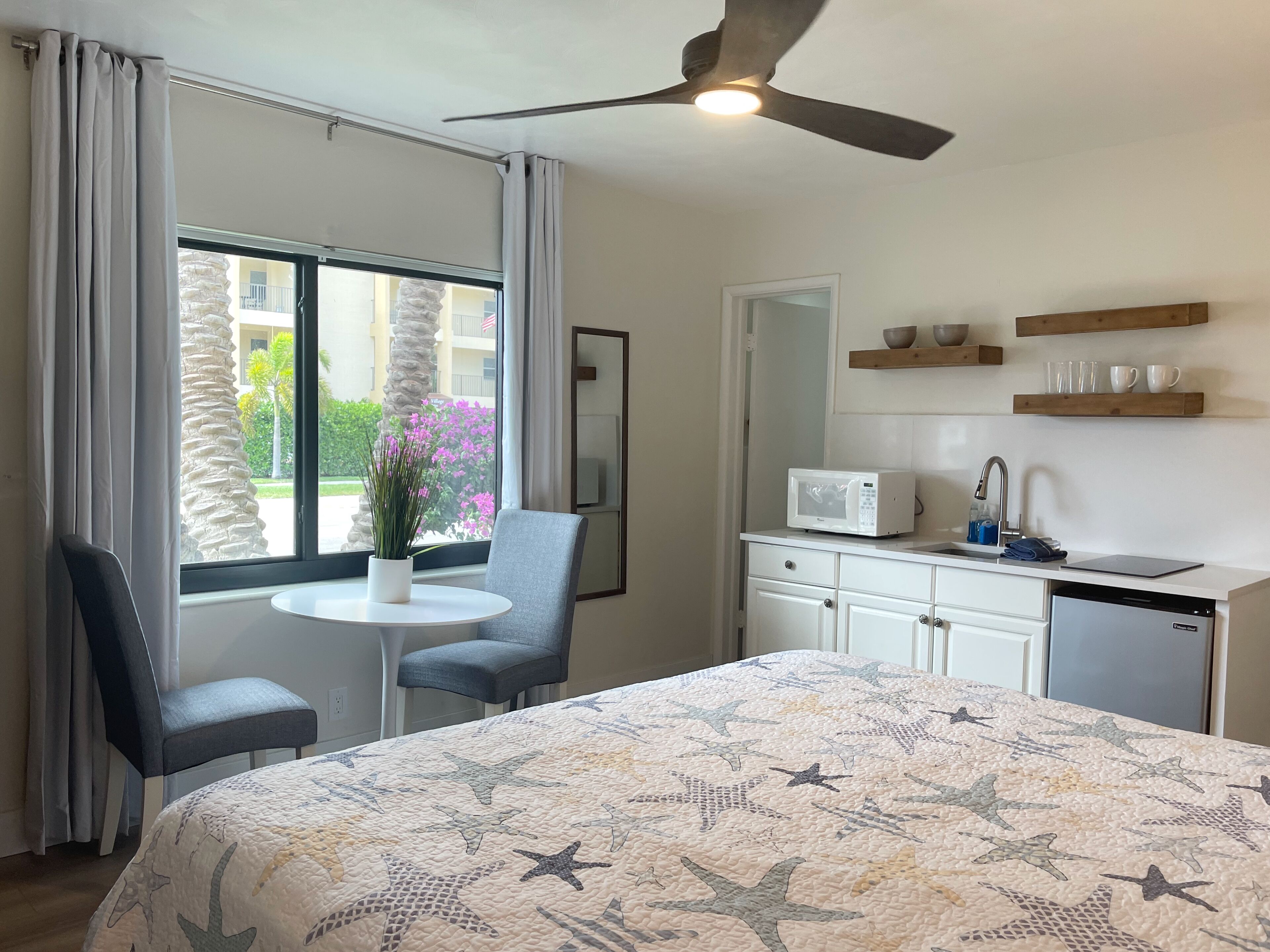 A hotel room with a bed, a desk, a chair, a small table with a potted plant, a microwave, a sink, and a ceiling fan.
