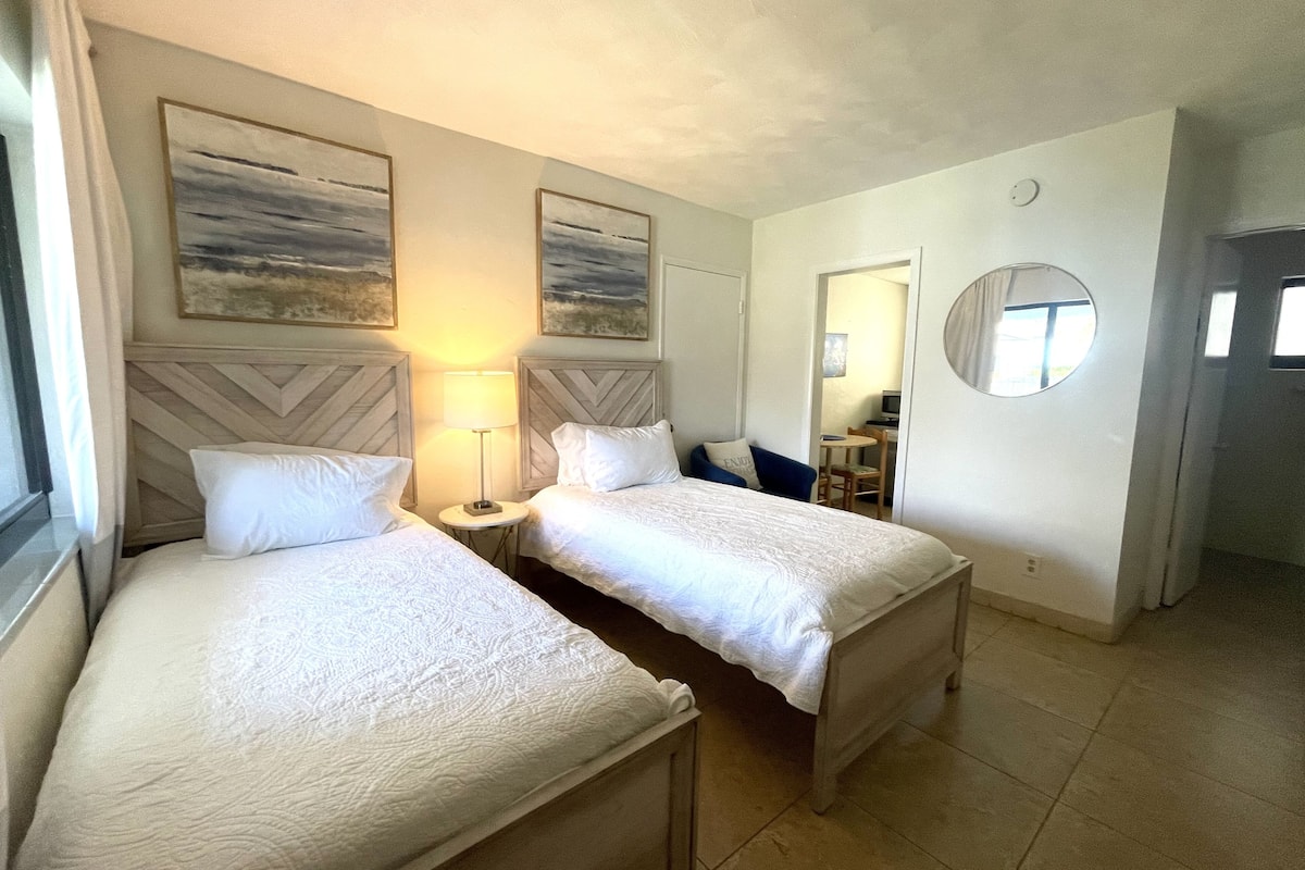 A hotel room with two beds, a small table, a lamp, a mirror, and artwork on the walls.