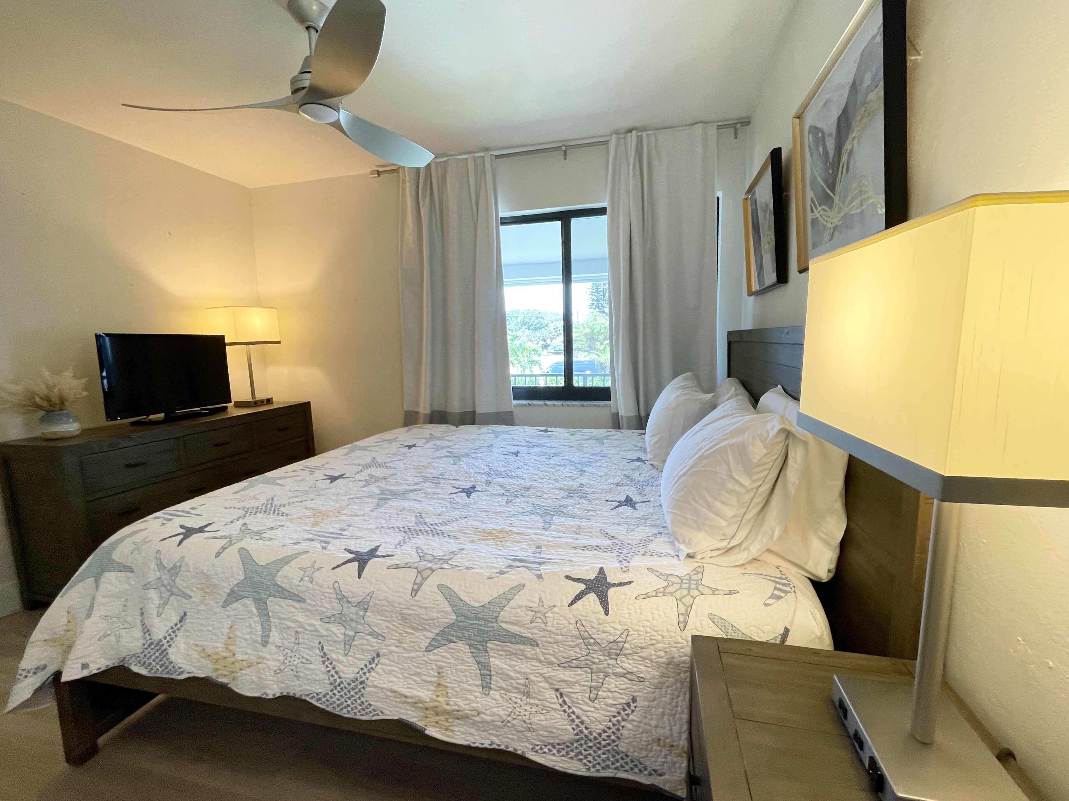 Un dormitorio con cama, televisor sobre una cómoda, ventilador de techo y ventana con cortinas.