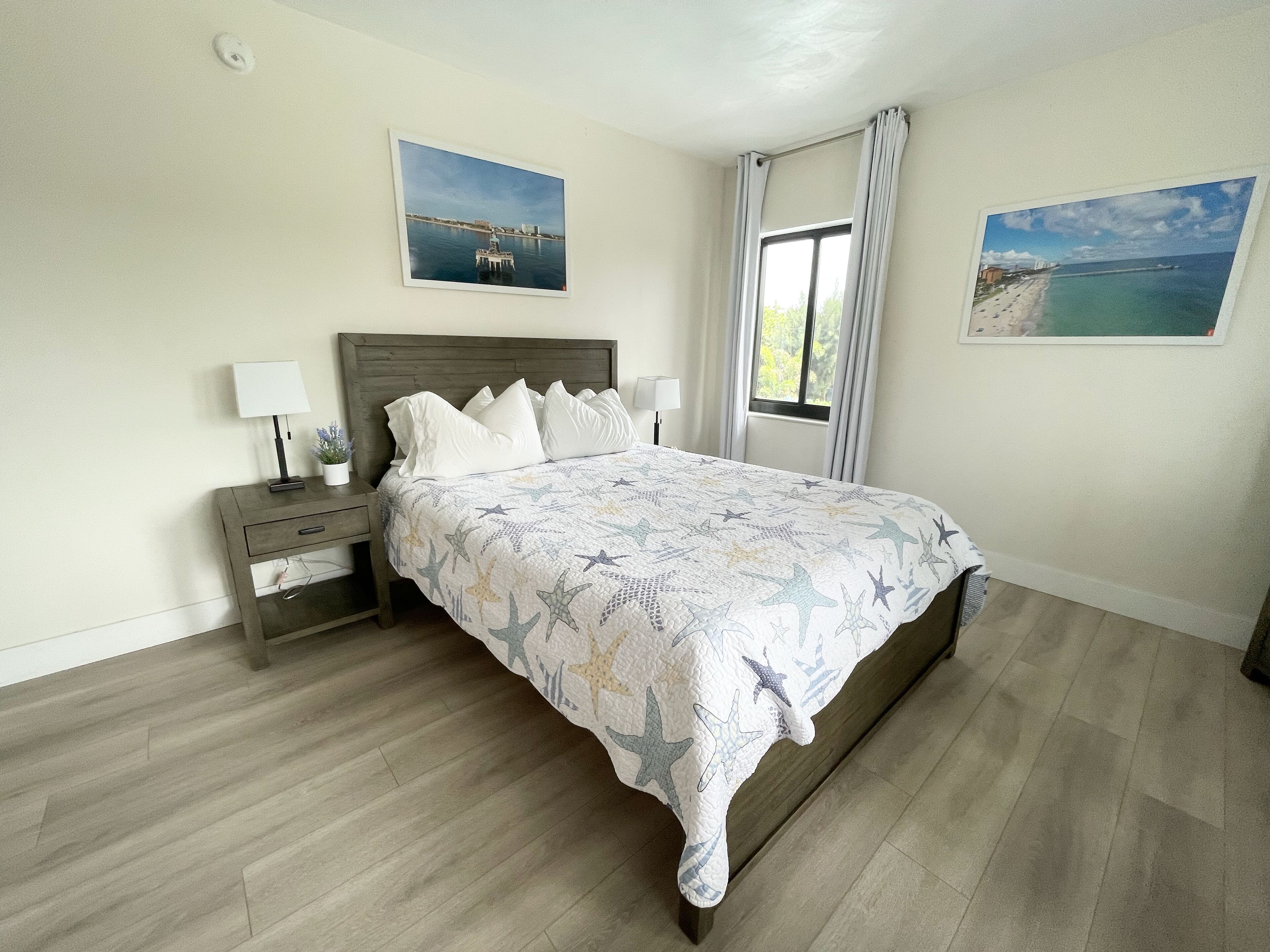 Un dormitorio con cama, mesitas de noche, una ventana con cortinas y dos cuadros en la pared.