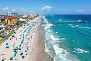 A beach with numerous umbrellas, a pier extending into the ocean, and buildings in the background. - Tropic Isle Beach Resort (Deerfield Beach)
