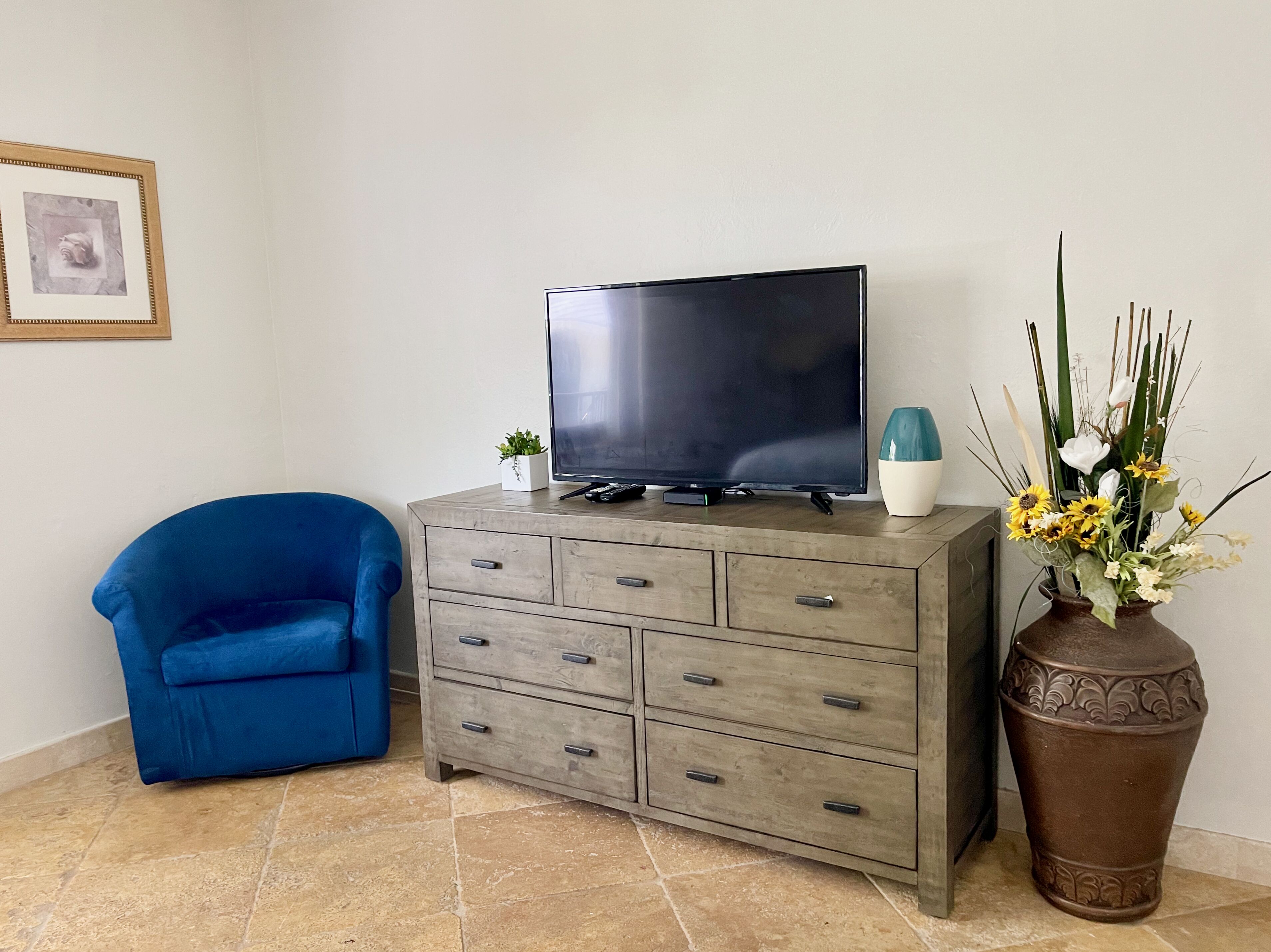 Een woonkamer met een televisie op een houten kast, een blauwe stoel en een decoratieve vaas met bloemen.