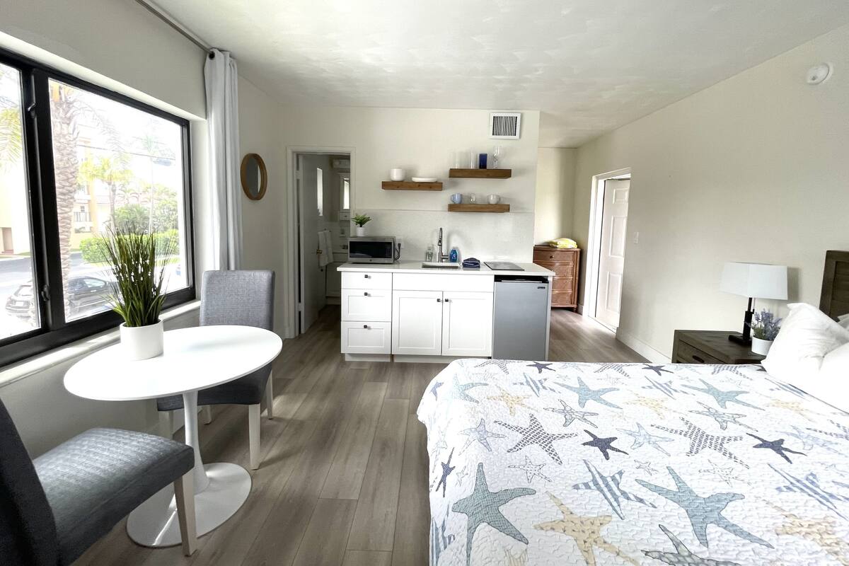 A hotel room with a bed, a small table with a chair, a kitchenette, and a window with a view of palm trees.