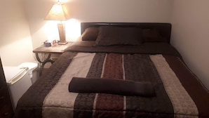 Een netjes opgemaakt bed met een gestreept dekbed en een kussen. Er staat een nachtkastje met een lamp en een klein doosje.