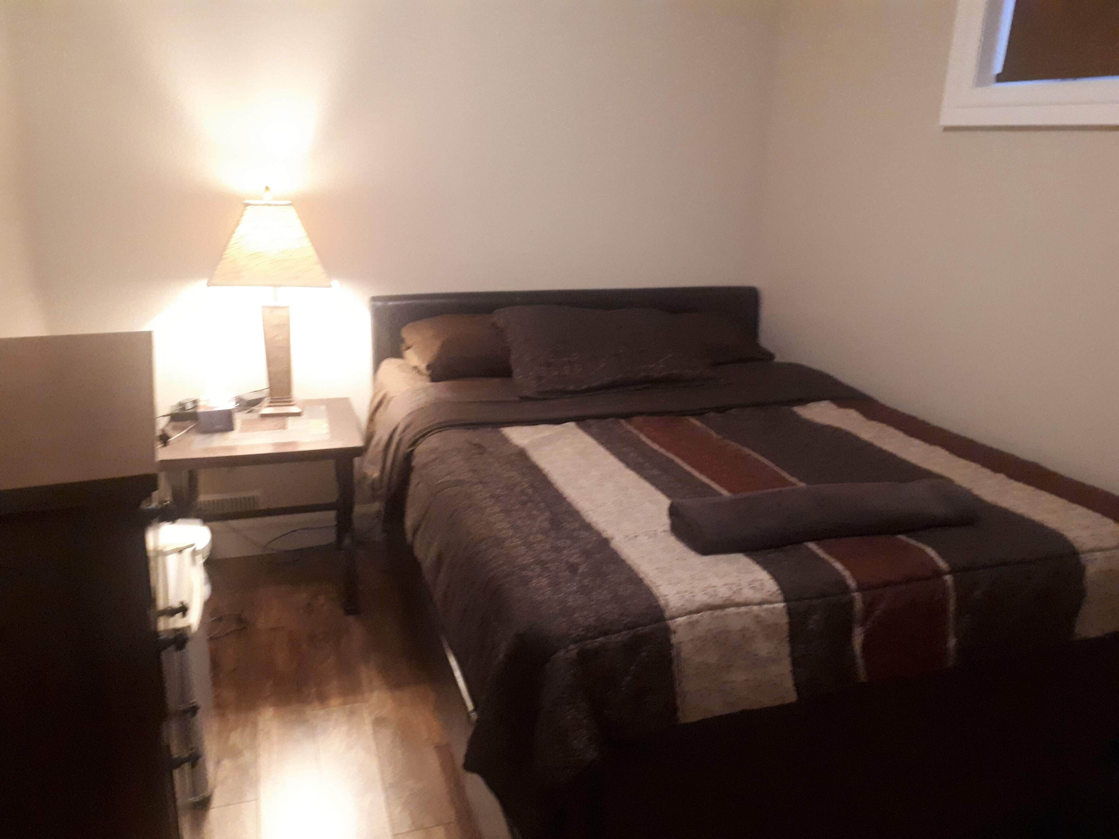Ein kleiner, schummrig beleuchteter Raum mit einem Einzelbett, einem Nachttisch mit Lampe und einem Fenster.