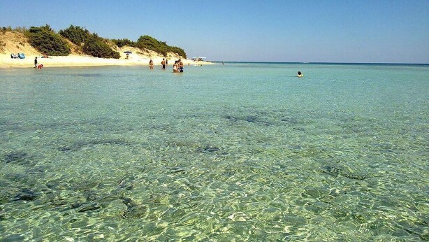 A beach with clear shallow waters, sandy shore, and people in the distance.
