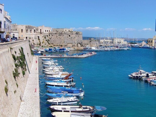 A harbor with numerous boats docked along a stone quay, a historic stone wall, and a clear blue sky.