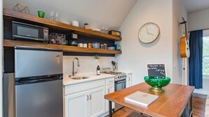 A kitchen with white cabinets, a stainless steel refrigerator, a microwave, and a wooden shelf above the sink. There's a green bowl on a wooden table, a clock on the wall, and a guitar leaning against the wall.