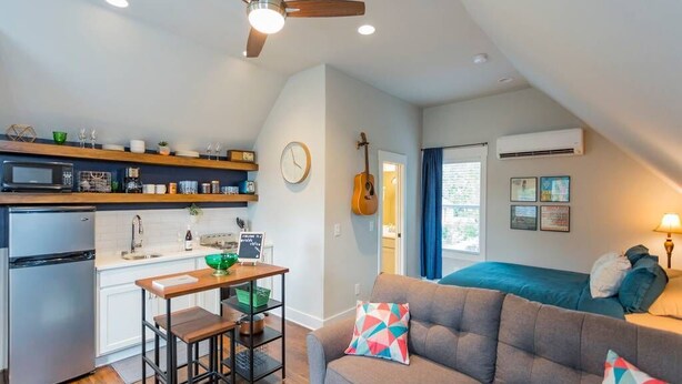 A compact living space with a kitchenette, a dining area, a sofa with colorful cushions, a guitar, and a bed with a blue headboard.