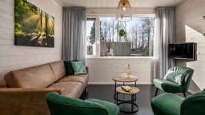 A cozy living room with a brown sofa, green armchairs, a small round table with a bottle and glasses, and a large window with a view of trees and a building. - Water Lodge in Schaijk by the Waterside (Schaijk)