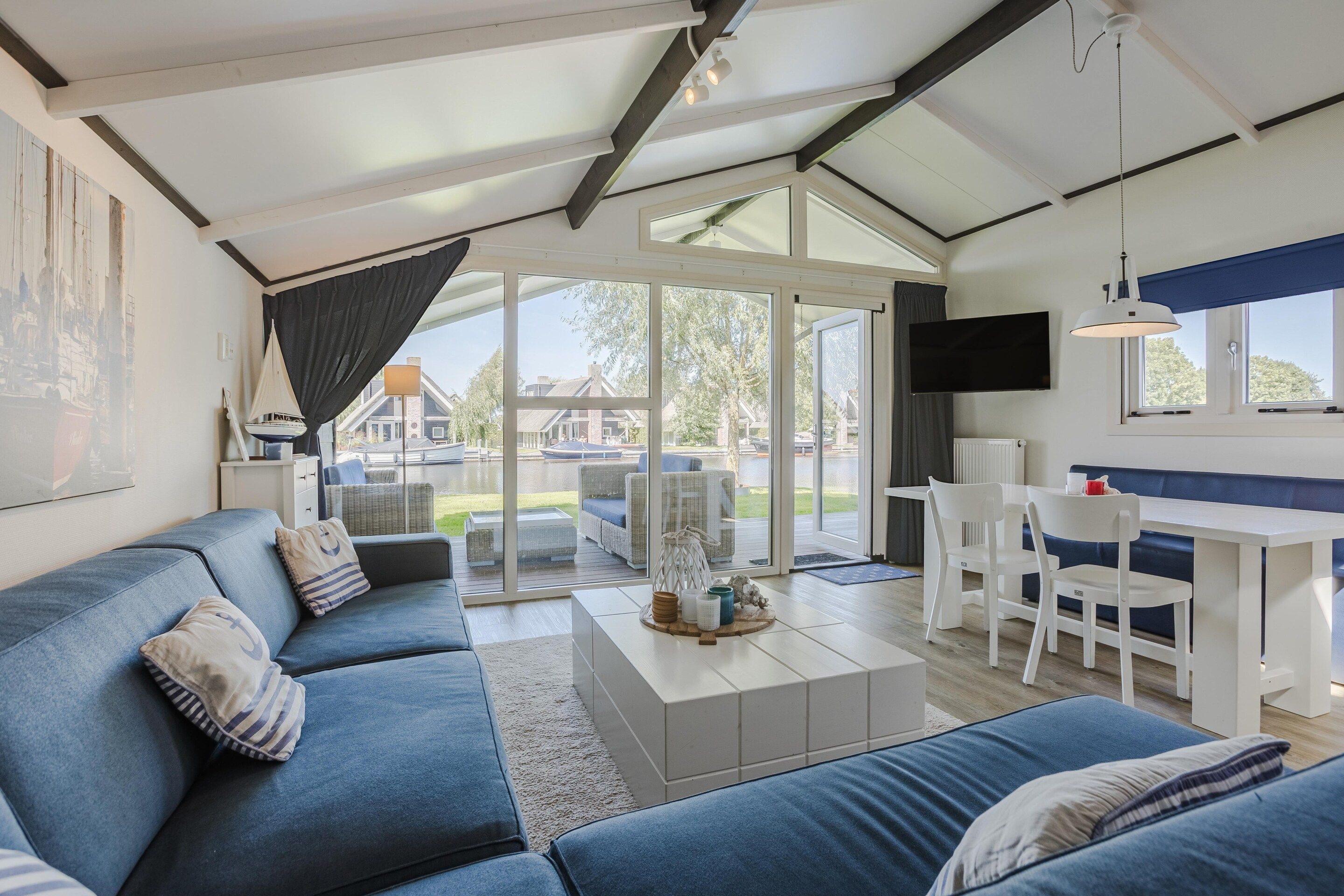 A modern living room with a blue sofa, a dining area with a white table and chairs, and large glass doors leading to a deck.