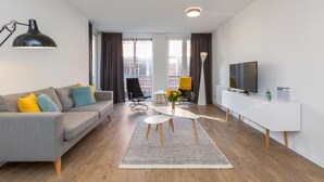A modern living room with a sofa, a coffee table, a TV, and a large window with curtains. - Modern Beach Apartment Zoutelande (Zoutelande)