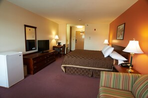 Una habitación de hotel con una cama, televisión, escritorio con lámpara, silla y refrigerador.