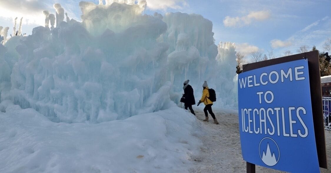A sign reading "WELCOME TO ICECASTLES" with a snowy landscape and two people walking.