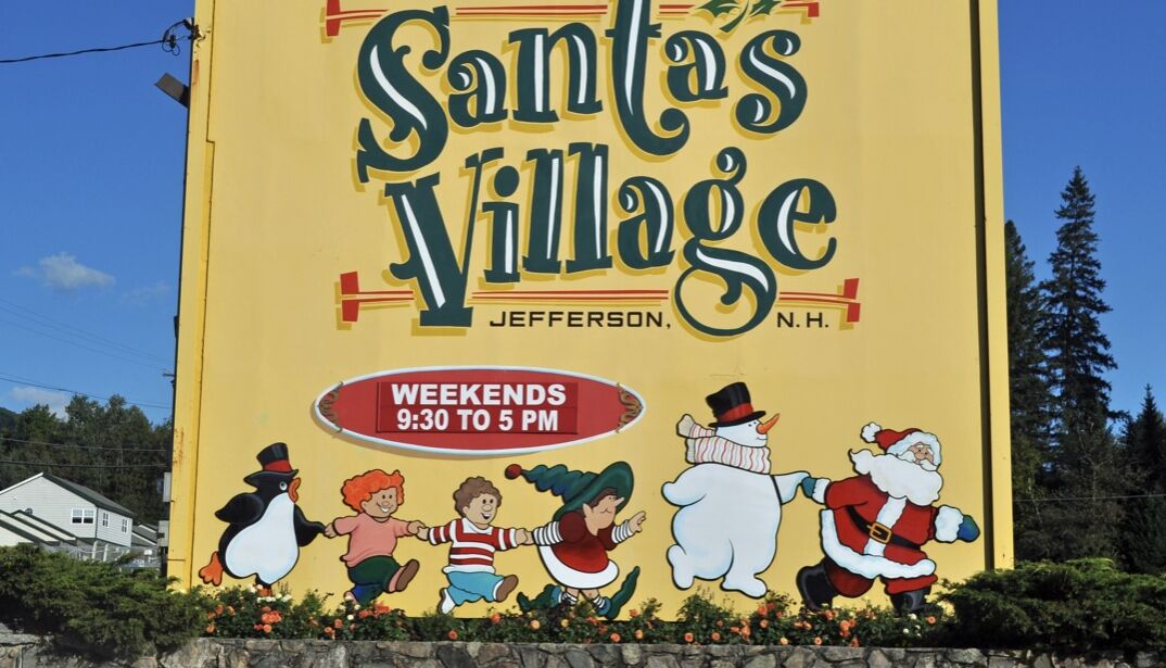 Um grande outdoor amarelo publicando 'Santa's Village' em Jefferson, NH, com uma ilustração festiva de crianças e Papai Noel.
