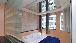 Quarto de hotel moderno com cama, divisória de vidro e grandes janelas com vista para a cidade.