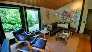 A modern living room with a sofa, a coffee table, and a chair. There is a large window with a view of greenery outside. - Bungalow in Waldbrunn (Waldbrunn)
