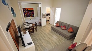 Un espacio habitable compacto con comedor, un sofá decorado con cojines y una pequeña cocina a la vista a través de una puerta abierta.