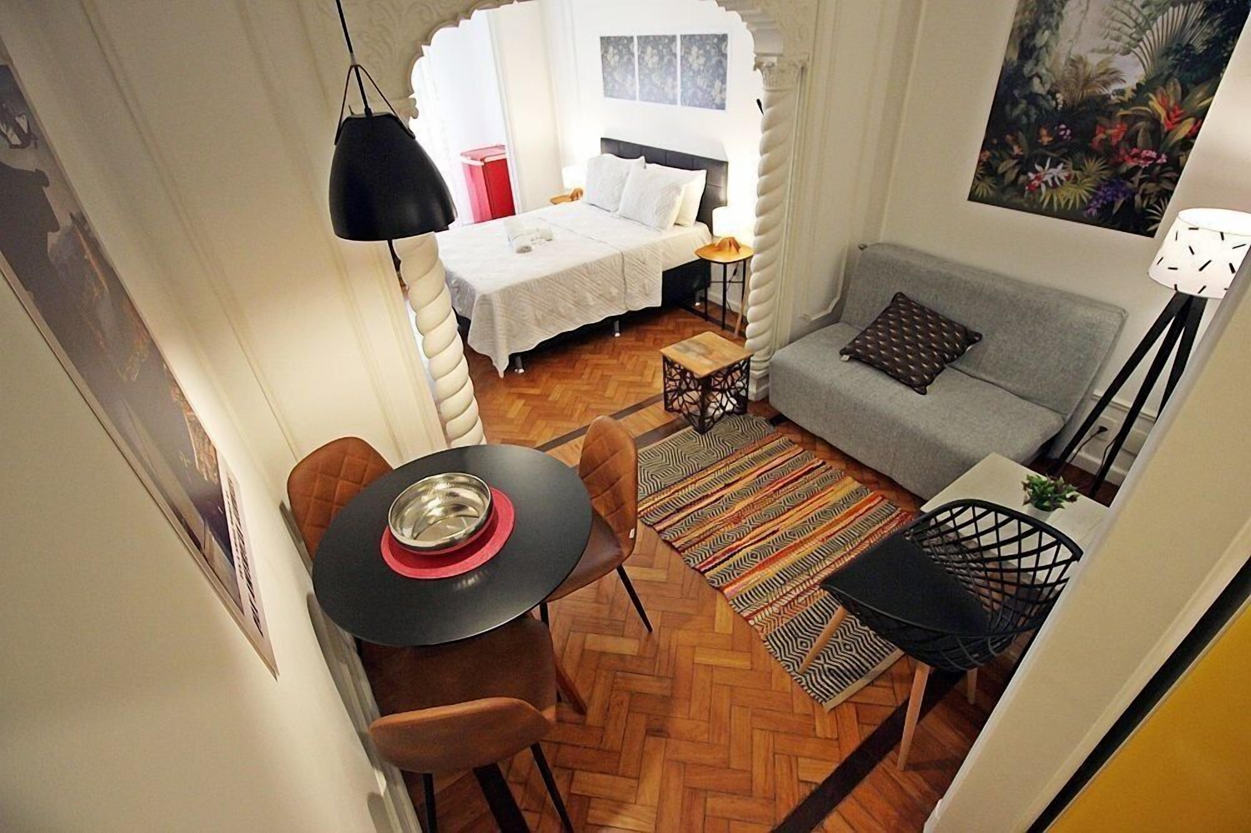 A compact room with a bed, a gray sofa, a dining table with chairs, and a small side table.