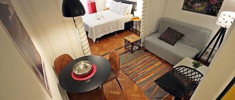 A compact room with a bed, a gray sofa, a dining table with chairs, and a small side table.