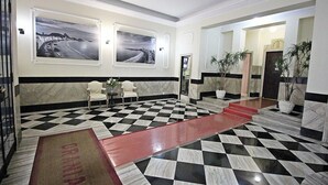 Un vestíbulo de hotel con suelo a cuadros, dos fotografías en blanco y negro enmarcadas, un mostrador de recepción y una escalera que conduce a otra zona.